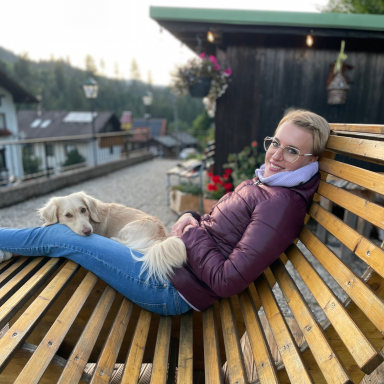 Eine gewölbte Holzliege auf welcher eine Frau mit blondem Haar und Steppjacke liegt. Auf hier ein kleiner amerikanischer langhaar Dackel. 
